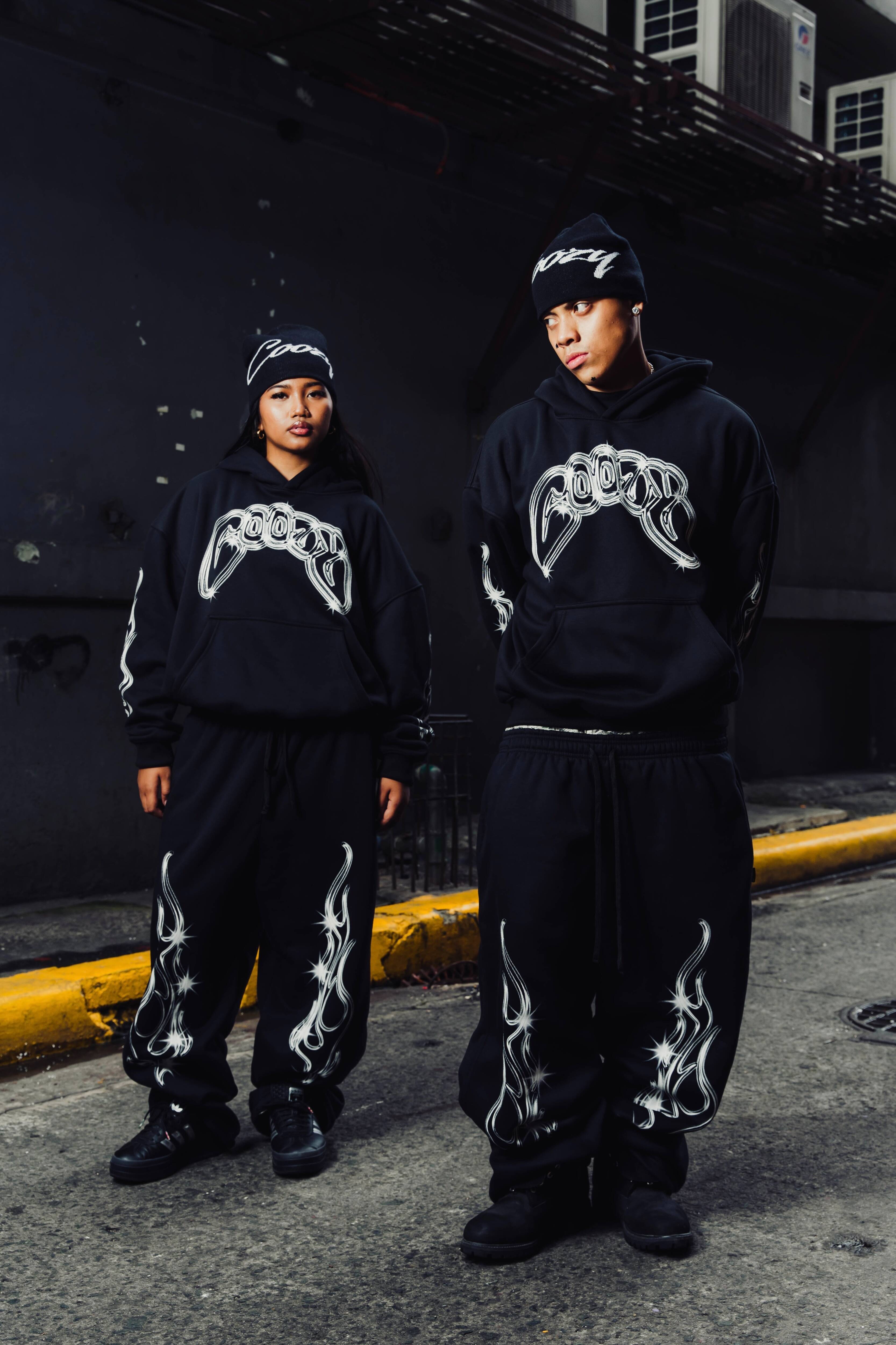 Two people wearing black hoodies with white designs standing on a street.