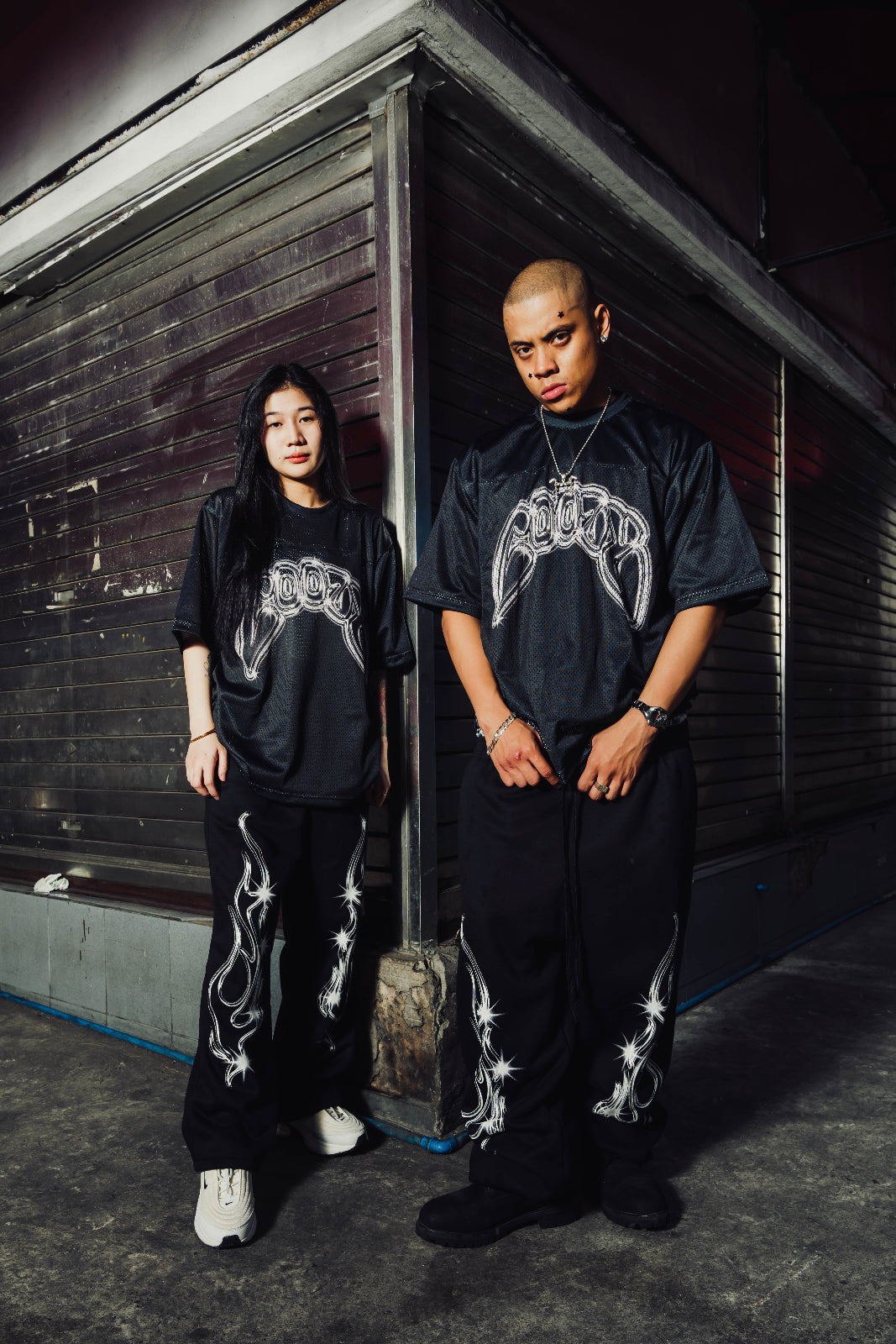 Two people wearing black outfits with white designs standing against a dark wall.